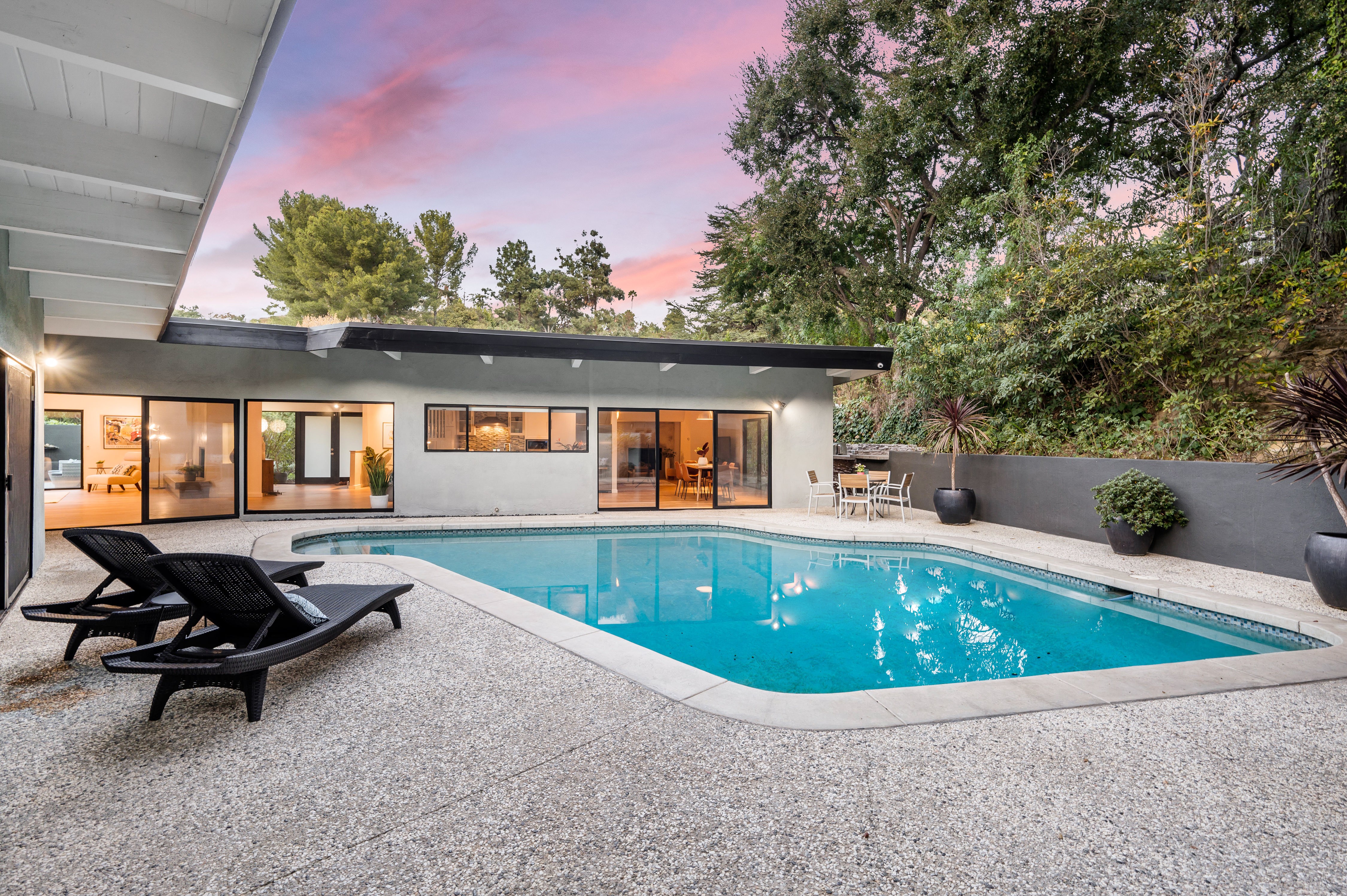 A modern home with a sleek pool area, featuring lounge chairs on a smooth, textured pool deck. The crystal-clear water of the pool complements the serene, green backdrop, making it an ideal outdoor space for relaxation and entertaining